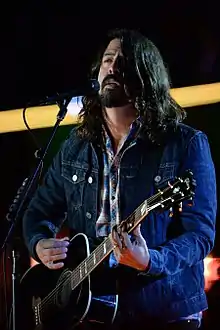 A middle-aged man with black hair and a beard sitting down playing a guitar.