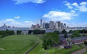 Aerial view of Governors Island in New York City on a sunny day. There is a lawn and some houses in the foreground, and skyscrapers in the background.